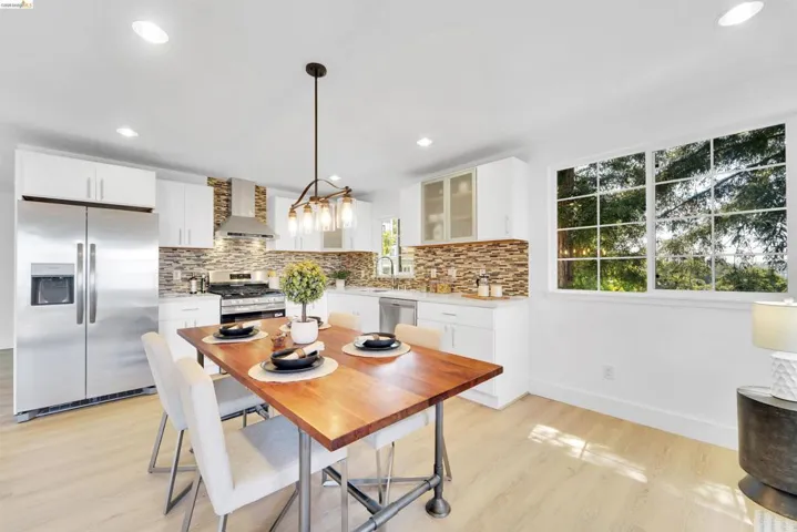 Open-concept kitchen and dining area featuring stainless steel appliances, white cabinetry, wood-finish flooring, a mosaic tile backsplash, and a large multi-pane window