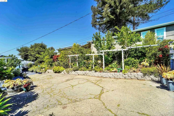 Expansive concrete driveway with tiered landscaping, a stone retaining wall, and a white pergola structure