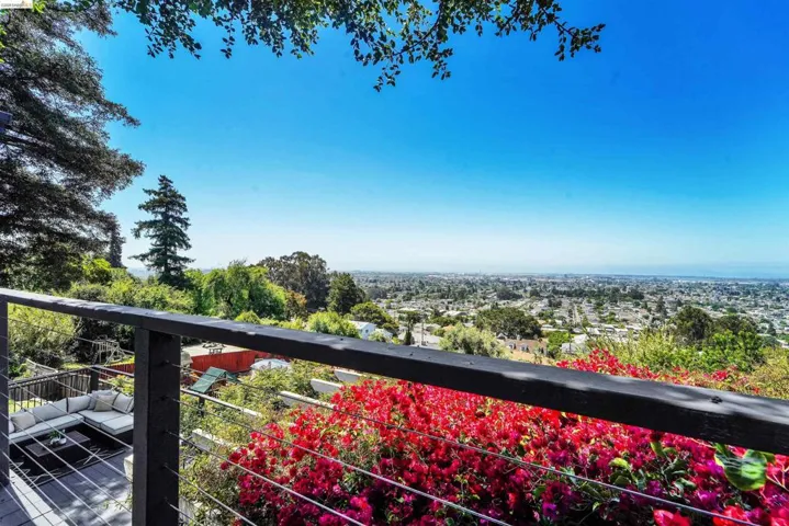 Expansive outdoor deck featuring a cable railing system, a lush garden with vibrant red flowering plants, and a panoramic cityscape extending to the horizon