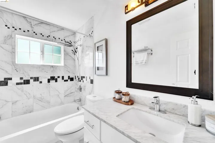 Bathroom featuring a marble-patterned tiled shower and tub combination with a horizontal black accent tile band, a vanity with a white rectangular sink and light-colored countertop, a dark-framed mirror, and a wall-mounted light fixture