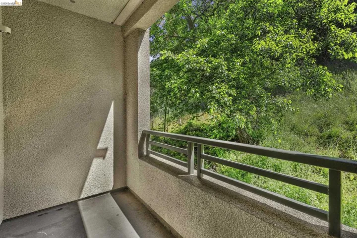 Private balcony with textured stucco walls, dark grey metal railing, and a verdant tree canopy backdrop