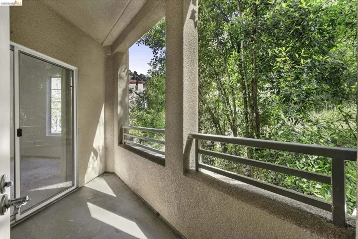 Balcony with a textured stucco exterior, sliding glass door entry, metal railing, and a verdant tree backdrop