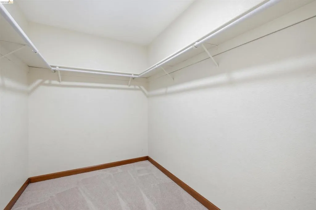 Walk-in closet featuring white wire shelving, hanging rods, light beige textured walls, light brown carpet, and wood-finish baseboards