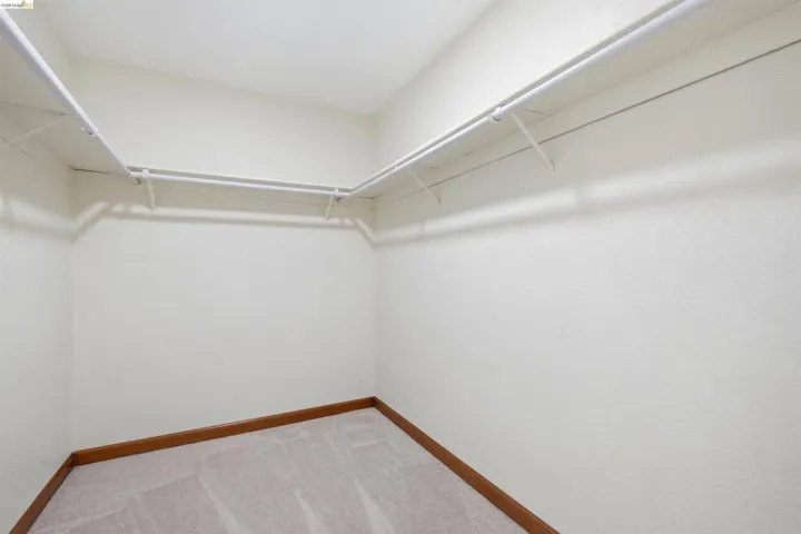 Walk-in closet featuring white wire shelving, hanging rods, light beige textured walls, light brown carpet, and wood-finish baseboards