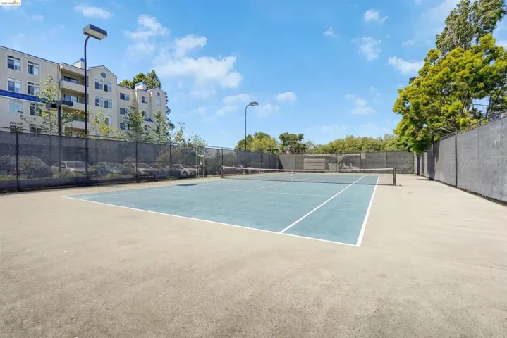 Tennis court featuring a blue playing surface with white line markings, surrounding concrete, perimeter fencing, and integrated light poles