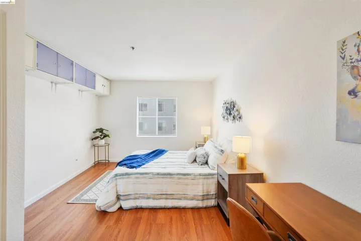 Bright bedroom featuring wood-finish flooring, a window with horizontal blinds, built-in overhead cabinetry, and white textured walls