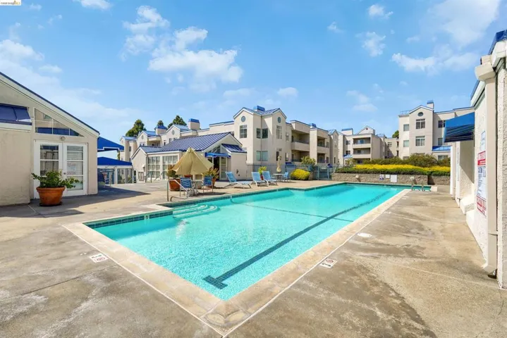 Outdoor swimming pool with concrete surround, framed by multi-story stucco buildings featuring blue roofs and awnings