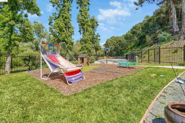 Expansive outdoor space featuring a manicured lawn, a dedicated play area with wood chip ground cover, a paved sports court, mature trees, and a black wrought-iron fence