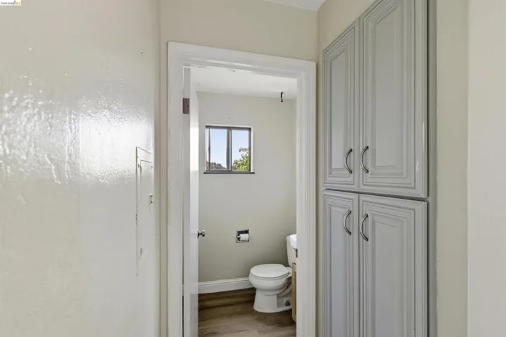 Bathroom featuring light gray shaker-style cabinetry with curved handles, wood-finish flooring, a window, and a white toilet