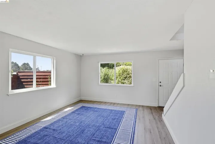 Open-concept living area featuring light wood-finish flooring, white walls, and two large windows providing natural illumination