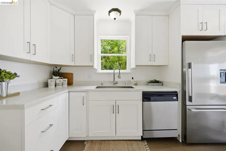 Bright kitchen featuring white shaker cabinetry, light countertops, stainless steel appliances, a window above the sink, and wood-finish flooring