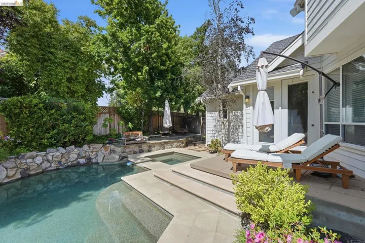 Outdoor pool and spa area featuring a rock wall, surrounding mature trees, and a spacious patio with multiple access points to the residence