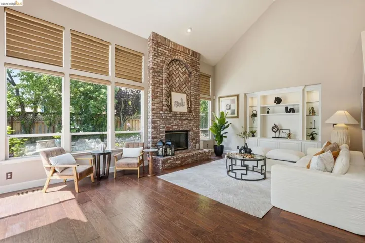Great room featuring a floor-to-ceiling brick fireplace with an arched hearth, expansive windows with integrated blinds, vaulted ceilings, built-in shelving, and wood-finish flooring