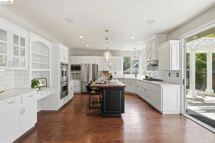 Spacious kitchen featuring wood-finish flooring, white cabinetry, a large center island with wood countertop, built-in stainless steel appliances, and a sliding glass door opening to an outdoor patio
