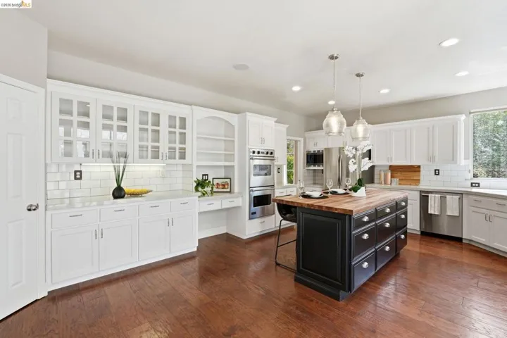 Spacious kitchen featuring a dark-toned island with a wood-finish countertop, white cabinetry, integrated stainless steel appliances, wood-finish flooring, and subway tile backsplash