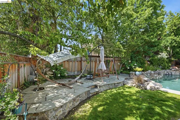 Landscaped backyard featuring a stone patio, mature trees, a manicured lawn, a privacy fence, and a rock-edged swimming pool