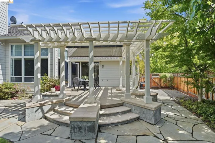Expansive flagstone patio featuring a white pergola, curved stone steps, a stone retaining wall, and a wood fence
