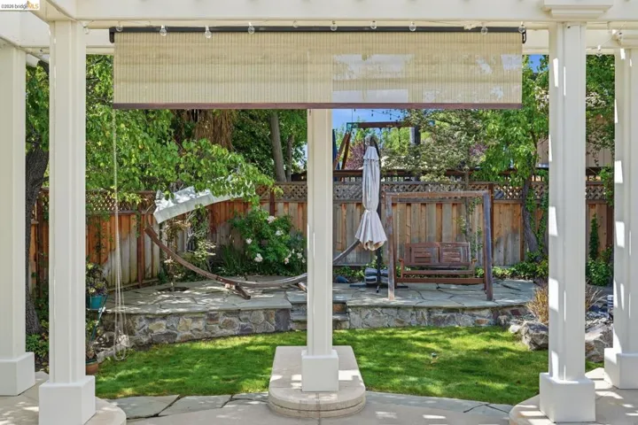 Backyard patio with a white pergola, textured privacy screen, natural flagstone patio, stone retaining wall, and wood fencing