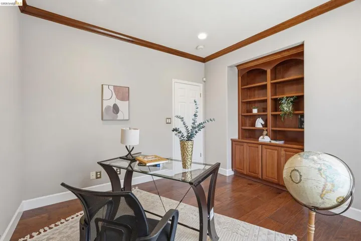 Dedicated office space featuring rich wood-finish flooring, an arched built-in bookcase with lower cabinetry, light gray wall tones, white baseboards, and elegant crown molding