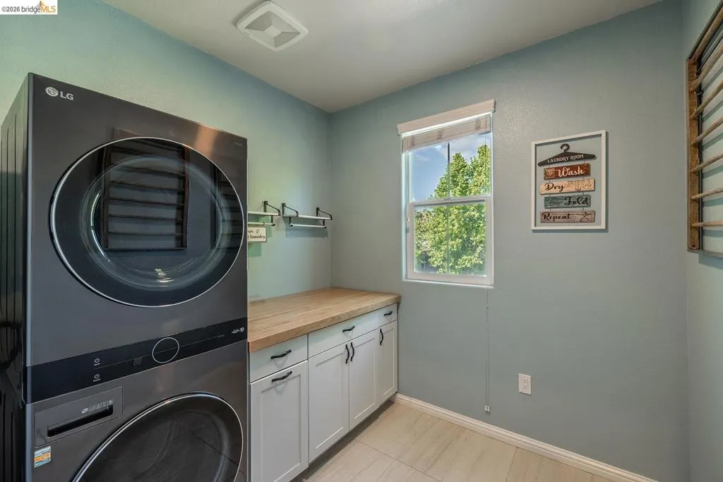 Dedicated laundry area featuring a stacked washer and dryer, wood-finish countertop, white base cabinetry with dark hardware, and a single window