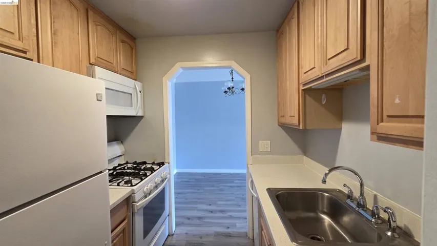 Galley kitchen featuring light wood cabinetry, white appliances, and a stainless steel sink