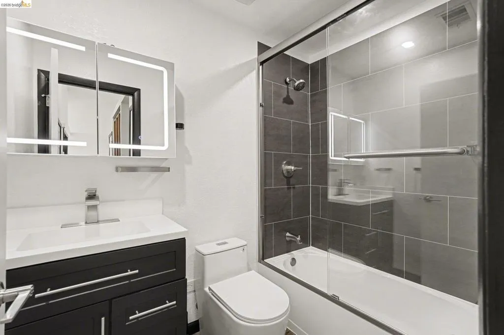 Contemporary bathroom featuring a dark wood-finish vanity with an integrated white sink, an illuminated mirrored medicine cabinet, a modern toilet, and a tiled shower-tub combination with a glass enclosure