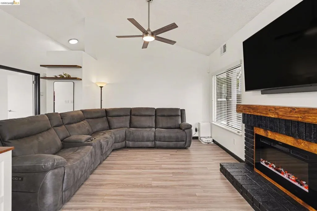 Living area featuring vaulted ceilings, wood-finish flooring, a modern ceiling fan, and a fireplace with a black brick surround and wood mantle