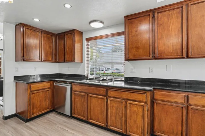 Kitchen featuring wood cabinetry, dark countertops, a stainless steel dishwasher, wood-finish flooring, and a window with blinds