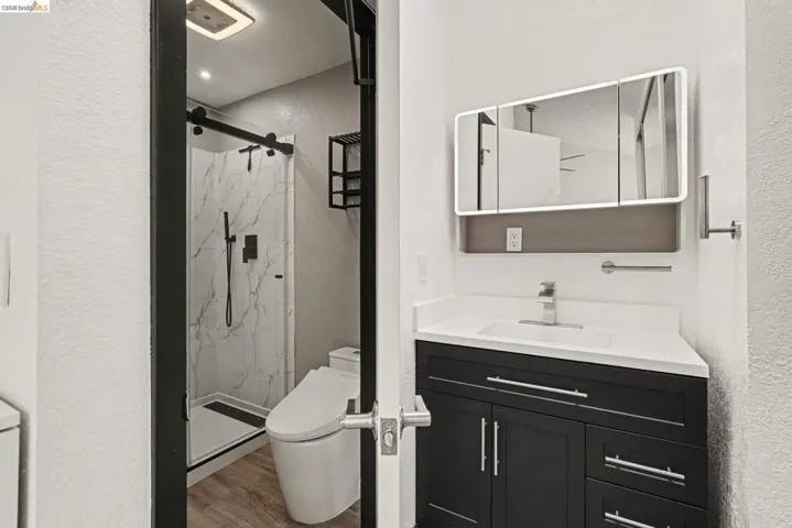 Contemporary bathroom featuring a dark wood vanity with a white countertop and rectangular sink, an illuminated mirrored medicine cabinet, a walk-in shower with a black-framed sliding glass door, white marble-look shower surround, and wood-finish flooring
