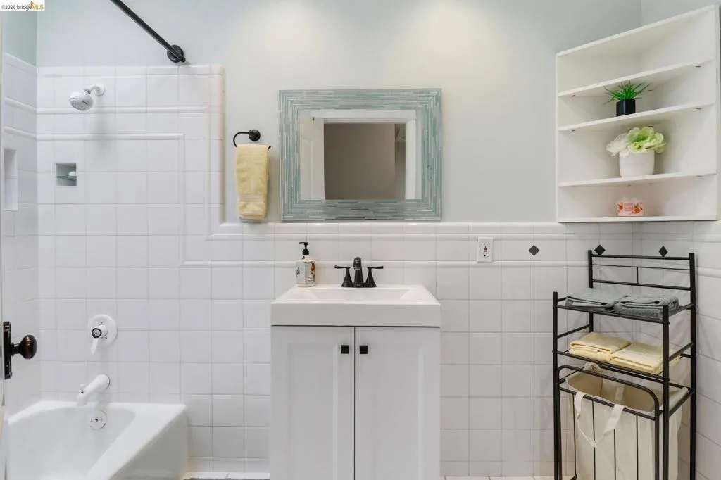 White tile bathroom featuring a bathtub with shower, white vanity with integrated sink, black faucet, and wall-mounted shelving