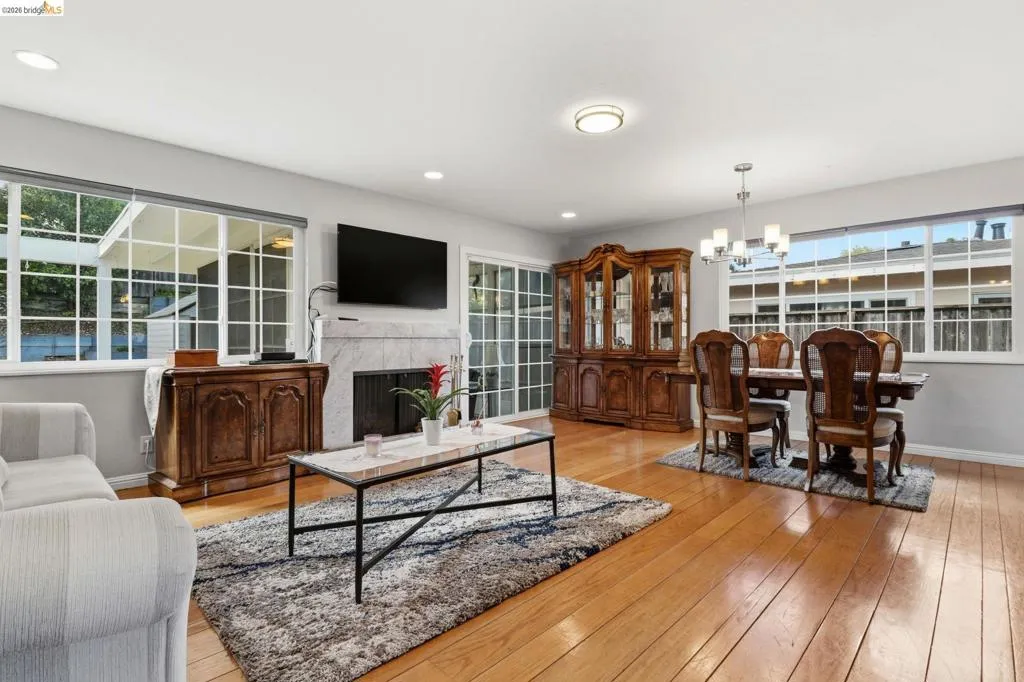 Open-concept living area featuring wood-finish flooring, a white stone fireplace surround, and multiple large windows with muntins