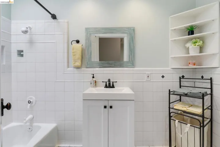 White tile bathroom featuring a bathtub with shower, white vanity with integrated sink, black faucet, and wall-mounted shelving