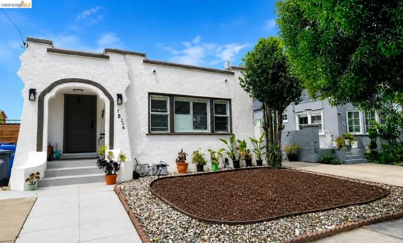 Stucco exterior featuring an arched entry with sconce lighting, dark trim accents, a front window with horizontal blinds, a landscaped front yard with a mulch bed and decorative rock, and a concrete pathway leading to the entry