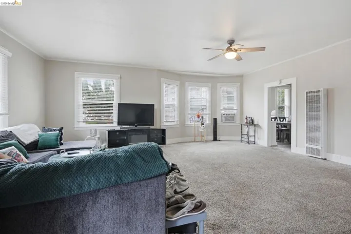 Spacious carpeted room featuring a bay window, ceiling fan, crown molding, a visible wall-mounted heater, and an open doorway to another area