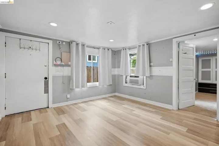 Spacious room featuring light wood-finish flooring, two windows, recessed ceiling lighting, a white painted door, and a light gray wall finish with a white chair rail