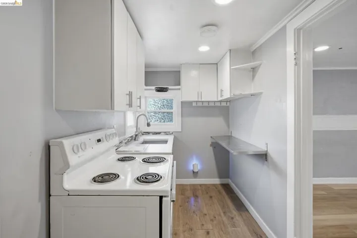 Compact kitchen featuring white cabinetry with brushed metal hardware, a white electric range, a stainless steel sink with a gooseneck faucet, wood-finish flooring, and a wall-mounted countertop extension