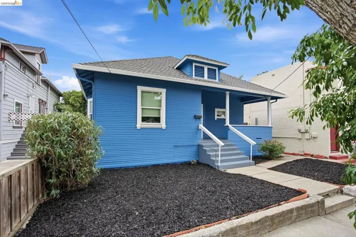 Exterior featuring vibrant blue siding, a covered porch with white columns, concrete steps with white railings, a front yard with dark ground cover, and a dormer window on the roof