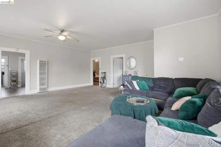 Spacious living area featuring neutral-toned walls, light-colored carpeting, a ceiling fan with integrated lighting, a wall-mounted heating unit, and white trim