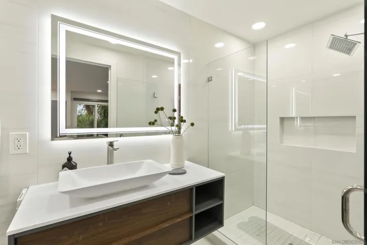 A sleek en-suite bath designed with clean modern lines, featuring an illuminated vanity mirror, floating wood cabinetry, and a glass enclosed shower that creates a refined guest experience.