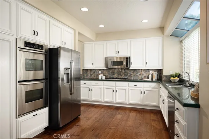 The kitchen features engineered wood flooring, Caesarstone countertops, stainless steel appliances, dual ovens, a tile backsplash and white cabinetry.