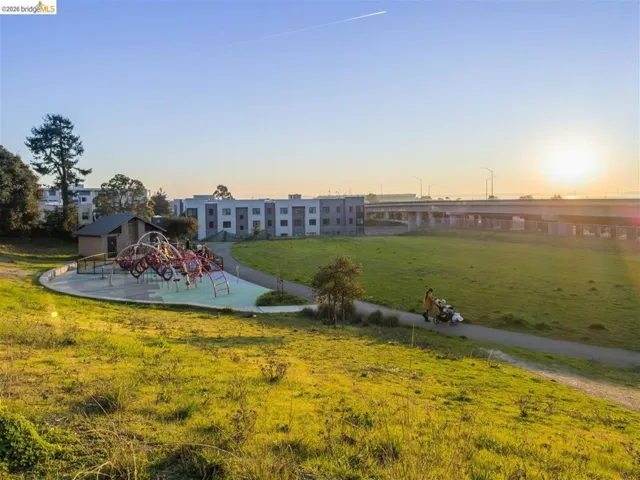 Expansive green space featuring a modern playground structure with red and white elements, a paved walking path, and a distinct building with a brown facade