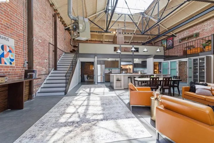 Expansive open-concept living space featuring exposed brick walls, industrial-style ceilings with exposed trusses and skylights, a built-in desk, and a staircase leading to an upper level