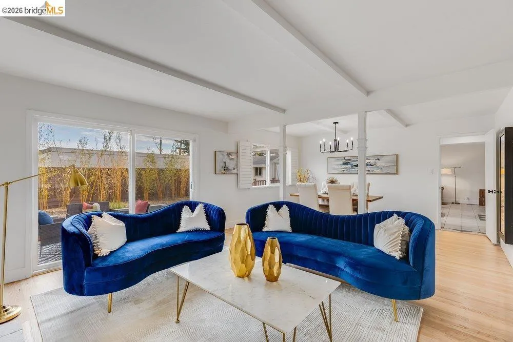 Bright living area featuring wood-finish flooring, white exposed beam ceiling, and a large sliding glass door opening to an outdoor patio area