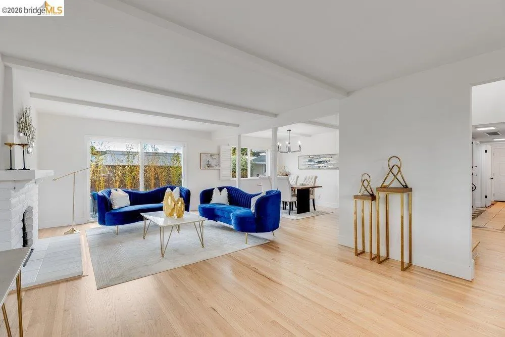 Bright living area featuring wood-finish flooring, a white brick fireplace with a tile hearth, and ceiling beams
