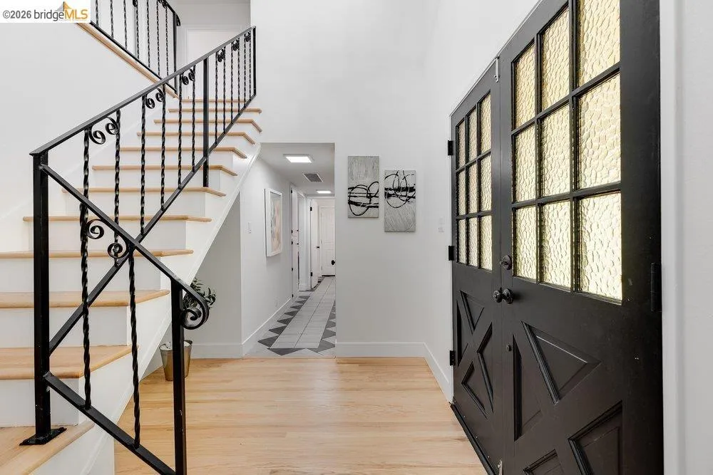 Entryway featuring a black double door with textured glass panels, light wood-finish flooring, a white staircase with black wrought iron railing, and a hallway with geometric tile flooring