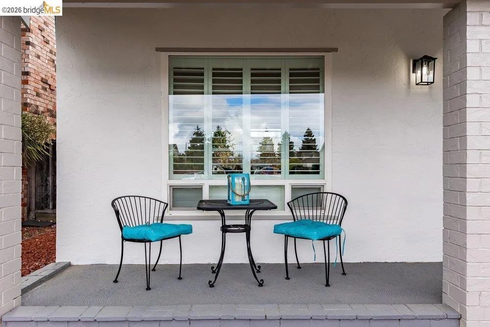 Covered porch featuring a window with interior shutters, textured stucco exterior, brick columns, an outdoor wall lantern, and a concrete patio surface