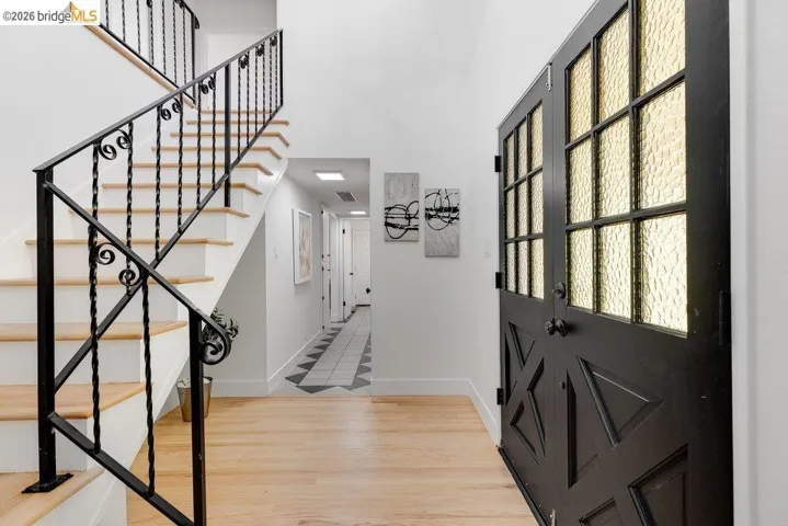 Entryway featuring a black double door with textured glass panels, light wood-finish flooring, a white staircase with black wrought iron railing, and a hallway with geometric tile flooring