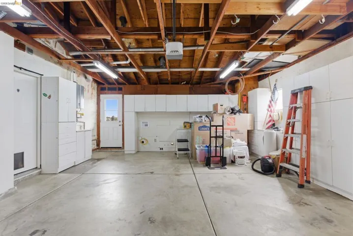 Spacious garage featuring exposed wood beam ceiling, concrete flooring, extensive built-in cabinetry, fluorescent lighting, and a rear access door with window