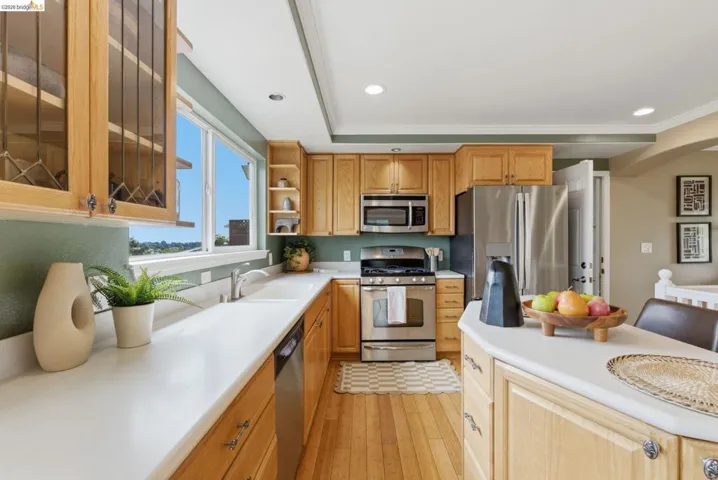 Open-concept kitchen featuring wood-finish cabinetry, stainless steel appliances, light-toned countertops, hardwood flooring, and a large window