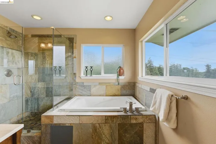 Recessed lighting illuminating a spacious bathroom featuring an integrated soaking tub, a separate glass-enclosed shower with stone tile, multiple windows providing natural light, and a brushed nickel towel bar